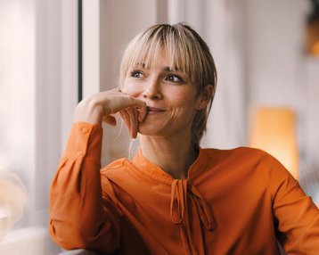 Eine Frau mit blonden Haaren und einem orangefarbenen Oberteil sitzt nachdenklich am Fenster und stützt ihr Kinn auf die Hand.