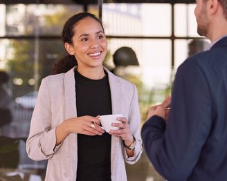 Eine Frau mit lockigem Haar trägt einen hellen Blazer und hält eine Tasse in der Hand, während sie mit einem Mann spricht. Im Hintergrund sind moderne Büromöbel sichtbar.
