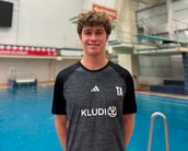Ein junger Mann steht vor einem Sprungbrett in einem Schwimmbad. Er trägt ein sportliches T-Shirt mit dem Aufdruck "TA" und "KLUDI". Im Hintergrund ist ein Sprungbrett sichtbar.