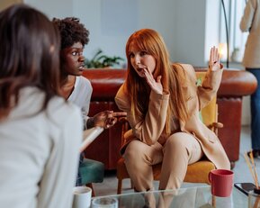 Drei Frauen sitzen in einem Café und unterhalten sich. Eine Frau mit langen roten Haaren gestikuliert lebhaft, während die anderen aufmerksam zuhören.