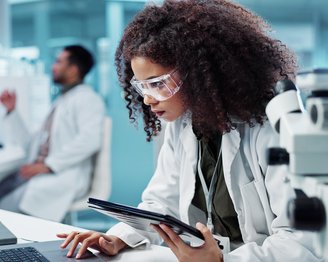 Eine Wissenschaftlerin in einem Labor arbeitet konzentriert an einem Laptop und hält ein Tablet in der Hand. Im Hintergrund ist ein Kollege zu sehen.