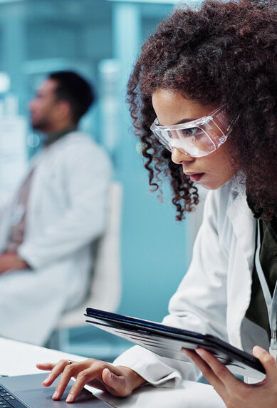 Eine Wissenschaftlerin in einem Labor arbeitet konzentriert an einem Laptop und hält ein Tablet in der Hand. Im Hintergrund ist ein Kollege zu sehen.
