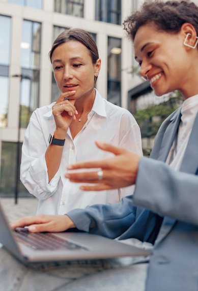 Zwei Frauen sitzen im Freien und schauen auf einen Laptop. Eine Frau zeigt auf den Bildschirm, während die andere nachdenklich wirkt. Beide tragen moderne Kleidung und sind in einem urbanen Umfeld.