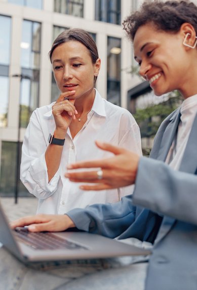 Zwei Frauen sitzen im Freien und schauen auf einen Laptop. Eine Frau zeigt auf den Bildschirm, während die andere nachdenklich wirkt. Beide tragen moderne Kleidung und sind in einem urbanen Umfeld.