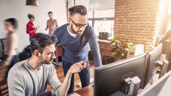 Zwei Männer diskutieren an einem Computer in einem modernen Büro. Einer zeigt auf den Bildschirm, während der andere aufmerksam zuhört. Im Hintergrund sind weitere Personen zu sehen, die an ihren eigenen Aufgaben arbeiten.