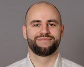 Ein Mann mit kurzem, dunklem Bart und Glatze trägt ein weißes Hemd. Er lächelt und schaut direkt in die Kamera. Der Hintergrund ist grau.