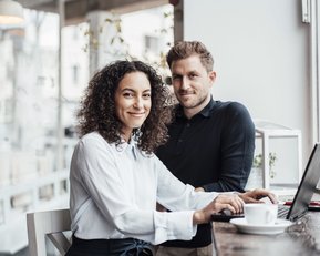 Ein Mann und eine Frau sitzen an einem Tisch in einem Café. Die Frau hat lockige Haare und trägt eine weiße Bluse, während der Mann in einem schwarzen Pullover gekleidet ist. Auf dem Tisch steht eine Tasse.
