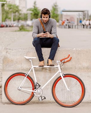 Ein Mann sitzt auf einer Betonmauer und schaut auf sein Smartphone. Neben ihm steht ein Fahrrad mit weißen Rahmen und orangefarbenen Reifen.