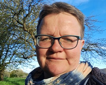 Eine Person mit Brille und einem bunten Schal steht im Freien, umgeben von Bäumen und grünem Gras. Der Himmel ist blau mit einigen Wolken.