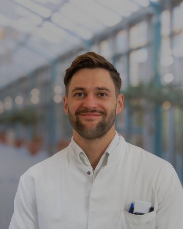 Ein Mann in einem weißen Kittel steht lächelnd in einem modernen, hellen Raum mit Pflanzen im Hintergrund.