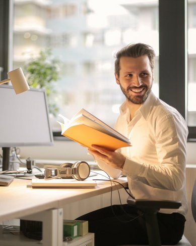 Ein Mann sitzt an einem Schreibtisch in einem modernen Büro, hält ein Buch in der Hand und lächelt. Auf dem Tisch befinden sich Computerbildschirme und ein Kopfhörer.