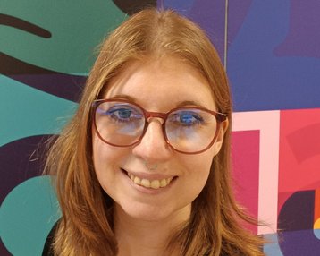 Eine Person mit langen, blonden Haaren und Brille lächelt in die Kamera. Im Hintergrund sind bunte geometrische Muster zu sehen.