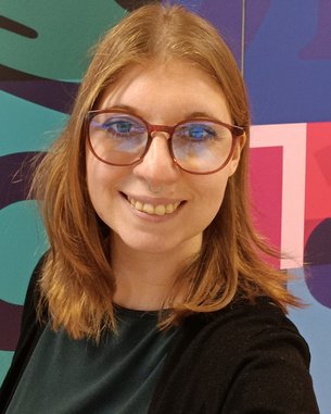 Eine Person mit langen, blonden Haaren und Brille lächelt in die Kamera. Im Hintergrund sind bunte geometrische Muster zu sehen.