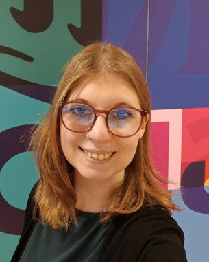 Eine Person mit langen, blonden Haaren und Brille lächelt in die Kamera. Im Hintergrund sind bunte geometrische Muster zu sehen.