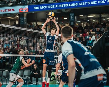 Ein Volleyballspieler hebt den Ball zum Zuspiel, während seine Teamkollegen und Gegner auf dem Spielfeld positioniert sind. Zuschauer im Hintergrund.