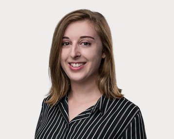 Eine junge Frau mit langen, lockigen Haaren trägt ein gestreiftes Hemd und lächelt in die Kamera. Der Hintergrund ist neutral.