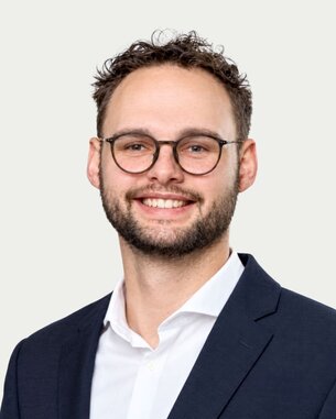 Ein junger Mann mit Brille und Bart lächelt, trägt einen Anzug und ein Hemd. Der Hintergrund zeigt unscharfe, helle Farben.