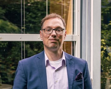 Ein Mann in einem Anzug steht entspannt vor einer Glaswand, umgeben von Pflanzen. Er trägt eine Brille und hat die Hände in den Taschen.