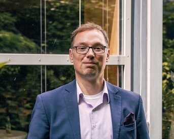 Ein Mann in einem Anzug steht entspannt vor einer Glaswand, umgeben von Pflanzen. Er trägt eine Brille und hat die Hände in den Taschen.