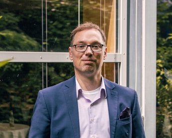 Ein Mann in einem Anzug steht entspannt vor einer Glaswand, umgeben von Pflanzen. Er trägt eine Brille und hat die Hände in den Taschen.
