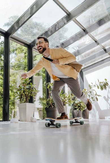 Mann in Anzug fährt auf einem Skateboard in einem modernen Raum mit großen Fenstern und Pflanzen.