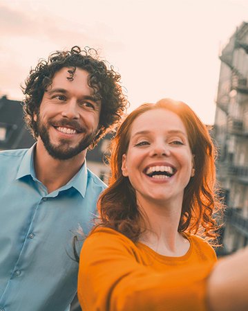 Ein Mann mit lockigem Haar und eine Frau mit langen, roten Haaren lächeln und machen ein Selfie vor einem urbanen Hintergrund. Beide tragen lässige Kleidung und wirken entspannt.