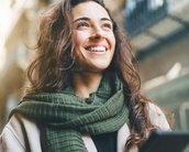 Junge Frau mit langen, lockigen Haaren und einem grünen Schal, die lächelt und ein Smartphone in der Hand hält. Sie steht in einer urbanen Umgebung mit unscharfem Hintergrund.