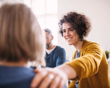 Resilienz stärken Eine lächelnde Frau mit lockigem Haar trägt einen gelben Pullover und legt ihre Hand auf die Schulter einer anderen Person. Im Hintergrund sind zwei weitere Personen zu sehen, die an einem Gespräch teilnehmen.