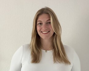 Eine lächelnde Frau mit langen, blonden Haaren trägt ein weißes Oberteil und steht vor einer hellen Wand.