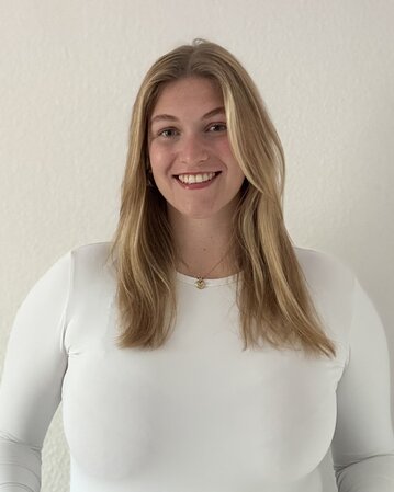 Eine lächelnde Frau mit langen, blonden Haaren trägt ein weißes Oberteil und steht vor einer hellen Wand.