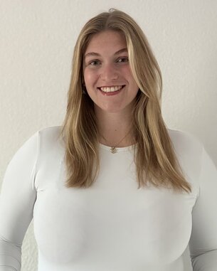 Eine lächelnde Frau mit langen, blonden Haaren trägt ein weißes Oberteil und steht vor einer hellen Wand.