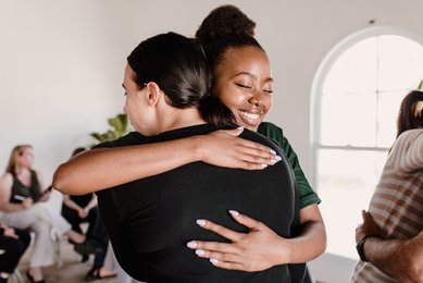 Zwei Frauen umarmen sich in einem hellen Raum. Eine Frau trägt ein schwarzes Oberteil, die andere ein grünes. Im Hintergrund sind weitere Personen zu sehen, die an einem Treffen teilnehmen.