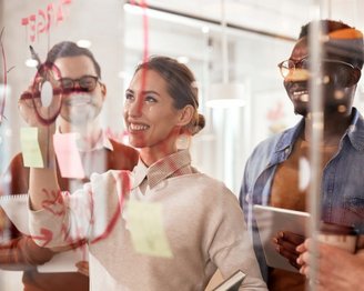 Eine Frau erklärt etwas an einem Glasboard, während drei Personen aufmerksam zuhören. Auf dem Board sind bunte Haftnotizen und Diagramme sichtbar.