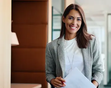 Eine lächelnde Frau in einem modernen Büro hält ein Dokument in der Hand. Sie trägt einen grauen Blazer und hat lange, braune Haare.