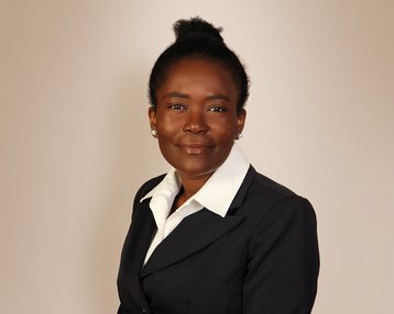 Eine Frau mit dunklem Haar, das zu einem Dutt gebunden ist, trägt einen schwarzen Blazer und ein weißes Hemd. Sie schaut direkt in die Kamera.