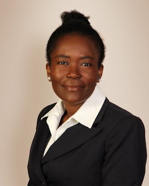 Eine Frau mit dunklem Haar, das zu einem Dutt gebunden ist, trägt einen schwarzen Blazer und ein weißes Hemd. Sie schaut direkt in die Kamera.