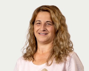 Eine Frau mit lockigem, braunem Haar lächelt freundlich in die Kamera. Sie trägt ein helles T-Shirt und hat einen neutralen Hintergrund.
