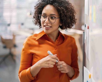 Eine Frau mit lockigem Haar und Brille steht vor einem Whiteboard und hält einen Marker, während sie spricht. Sie trägt ein oranges Hemd.