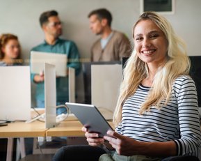 Eine lächelnde Frau mit langen, blonden Haaren hält ein Tablet in einem modernen Büro, während im Hintergrund zwei Männer und eine Frau sprechen.