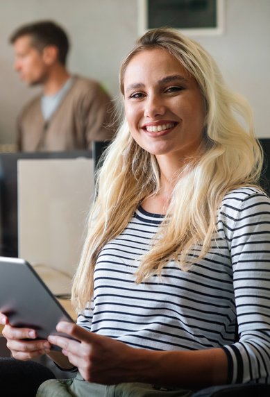 Eine lächelnde Frau mit langen, blonden Haaren hält ein Tablet in einem modernen Büro, während im Hintergrund zwei Männer und eine Frau sprechen.