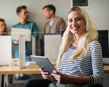 Eine lächelnde Frau mit langen, blonden Haaren hält ein Tablet in einem modernen Büro, während im Hintergrund zwei Männer und eine Frau sprechen.