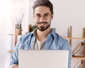 Ein Mann mit Brille lächelt, während er einen Laptop in den Händen hält. Er trägt ein hellblaues Hemd und steht in einem hellen Raum mit Regalen im Hintergrund.