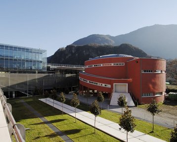 Moderne Architektur mit einem roten Rundbau und einem gläsernen Gebäude, umgeben von einer gepflegten Grünfläche und Bäumen, vor einer bergigen Kulisse.