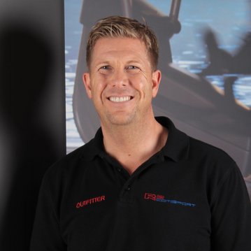 Ein Mann mit kurzem, blondem Haar trägt ein schwarzes Poloshirt mit dem Aufdruck "OUTFITTER". Er steht vor einem verschwommenen Hintergrund, der Wasser und Boote zeigt.