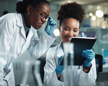 Zwei Wissenschaftlerinnen in Laborkitteln betrachten gemeinsam ein Tablet und diskutieren Ergebnisse in einem modernen Labor.