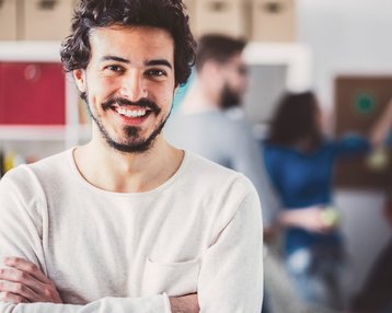 Ein lächelnder Mann mit lockigem Haar steht im Vordergrund, während Menschen im Hintergrund in einem kreativen Raum interagieren.