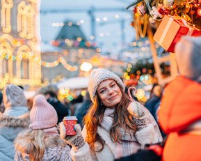Eine Frau mit einer Tasse in der Hand steht in einer belebten Weihnachtsmarkt-Szene, umgeben von festlich beleuchteten Ständen und Menschen.