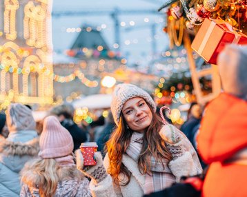 Eine Frau mit einer Tasse in der Hand steht in einer belebten Weihnachtsmarkt-Szene, umgeben von festlich beleuchteten Ständen und Menschen.