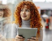 Eine junge Frau mit lockigem, rotem Haar hält ein Tablet in der Hand und schaut nachdenklich. Sie sitzt in einem modernen, hellen Raum mit großen Fenstern.