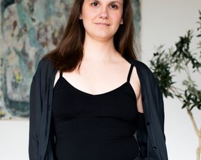 Eine Person mit langen, braunen Haaren trägt ein schwarzes Top und eine schwarze Jacke, steht vor einem abstrakten Kunstwerk und einer Pflanze.